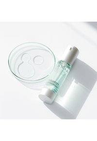Concentrato detergente in una bottiglia trasparente con un tappo argentato, posizionato accanto a una ciotola di vetro trasparente contenente gocce di liquido.