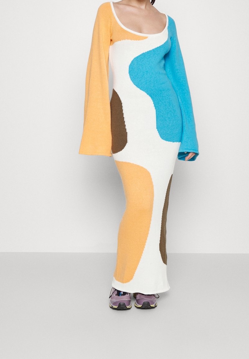 Femme portant une longue robe en maille multicolore avec des formes abstraites orange, bleues, marron et blanches, et des baskets violettes.