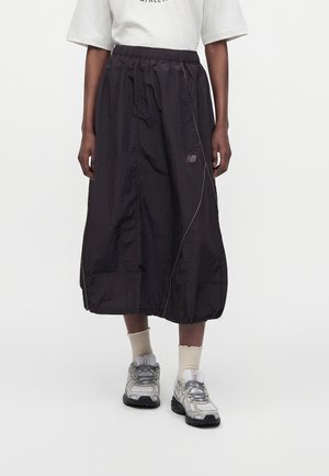 Personne portant une jupe noire mi-longue avec une taille élastique, des baskets blanches, des chaussettes beiges et un top blanc.
