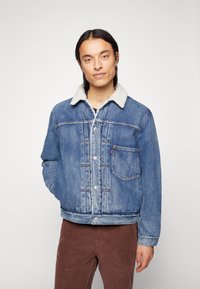 Leren denim jack in lichtblauw met een sherpa-kraag, zilveren knopen en een klassiek zakdesign. Draagt over een zwart shirt en bruine corduroy broek.