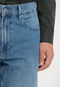 Jeans de mezclilla azul claro con un corte clásico, costuras visibles, bolsillos frontales y un cierre de botón metálico en la cintura.