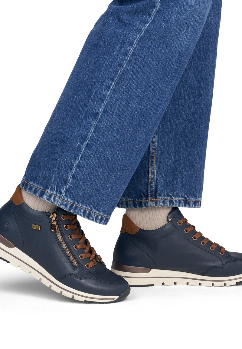 Personne portant un jean bleu et des chaussettes beiges avec des baskets en cuir bleu marine aux lacets et accents marron, debout sur un fond blanc.
