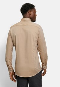 Man wearing beige long-sleeve buttoned shirt and dark trousers, facing away against a plain light background.