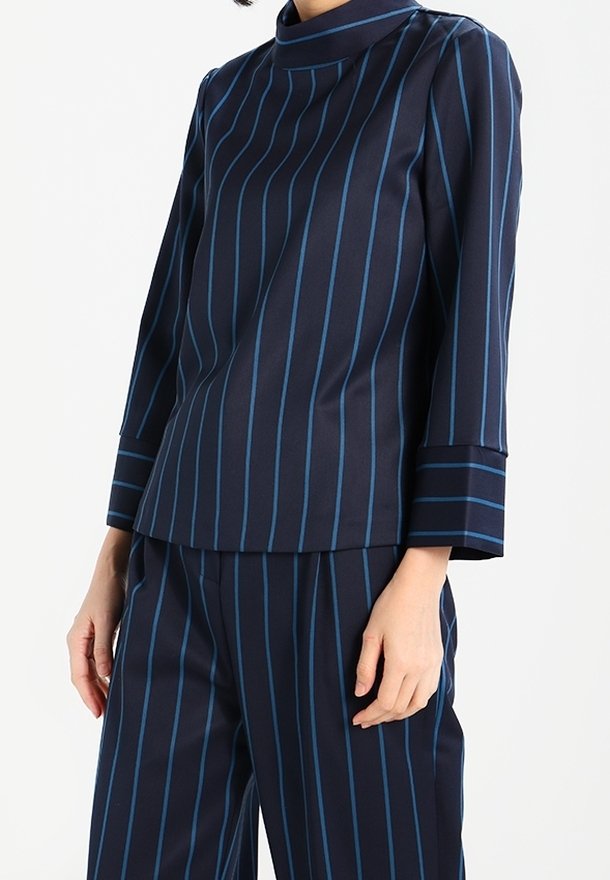 Femme portant un haut à manches longues bleu marine et un pantalon avec des rayures verticales bleu clair, debout devant un fond clair uni.
