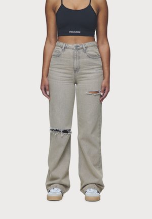 Mujer con vaqueros anchos beige rasgados y un top corto negro con logotipos visibles de "PEGADOR" en ambas prendas.
