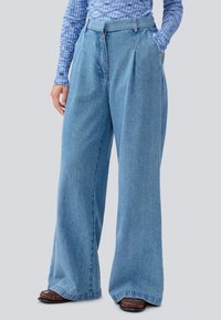 Lyseblå denim vid bukse med frontpress, to sidelommer og høy midje. Materialet har en myk tekstur med en svakt falmet finish.