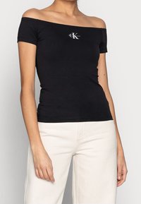 Czarny t-shirt z odkrytymi ramionami, wykonany z bawełny, z małym białym logo na środku klatki piersiowej, w zestawieniu z jasnymi spodniami.