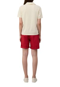 Polo en coton crème associé à un short rouge. Les shorts présentent des ourlets roulés. Modèle portant des chaussures claires, vue de dos.