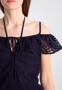 Top azul marino de encaje con diseño sin mangas, mangas con volantes, cordón ajustable y patrones florales texturizados en toda la prenda.