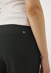 Černé elastické kalhoty s čistým designem, se zadní kapsou a malým kovovým logem. Texturovaný top ve světlé barvě.