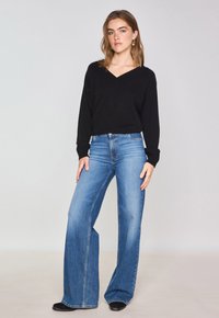 Un pull en maille noir avec un col en V associé à un jean large bleu clair. La tenue comprend des manches longues et une coupe décontractée. Des bottines noires sont visibles.