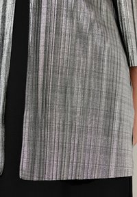 Glanzende, geplooide grijze stoffen jas met een metallic uitstraling, met een zwarte sectie eronder. Vlotte, reflecterende afwerking met verticale lijnen.