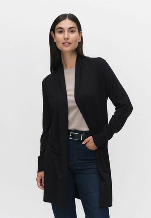 Cardigan noir ouvert à l'avant en tissu doux, avec des manches longues et une coupe décontractée, associé à un haut beige clair et à un jean foncé.