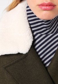 Abrigo verde oliva con un suave cuello blanco de piel, combinado con un suéter de cuello alto a rayas azul marino y blanco. La tela parece suave y con textura.