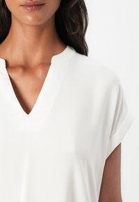 Weiße Bluse mit kurzen Ärmeln und V-Ausschnitt, die eine glatte Textur und subtilen Glanz aufweist. Der Stoff wirkt leicht und luftig.