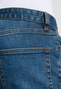 Pantalones vaqueros de mezclilla azul con el bolsillo trasero visible con costuras naranjas y un lazo para el cinturón, usados con una camisa blanca metida por dentro.