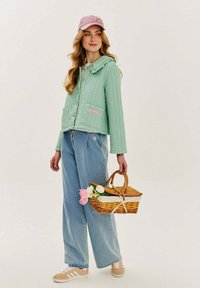 Mäta zelený prešívaný kabát s kapucňou a gombíkovým zapínaním, kombinovaný s svetlomodrými širokými džínami a béžovými teniskami. Drží prútený kôš.