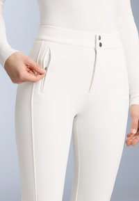 Pantaloni albi elasticizați cu o talie înaltă, cu fermoare laterale și două nasturi la talie, prezentați cu o mână care ajustează fermoarul.