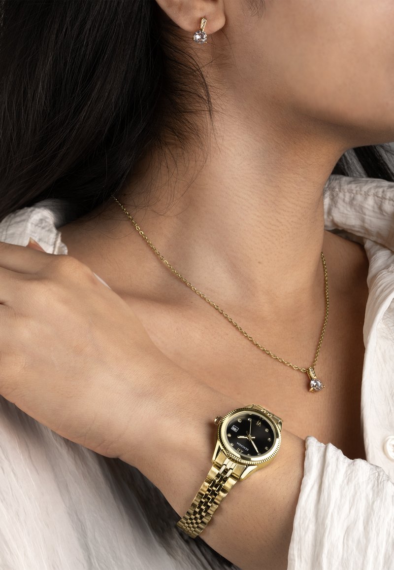 Montre en or avec cadran noir et bracelet en métal, accompagnée d'une chaîne en or avec un pendentif en cristal et des boucles d'oreilles clous assorties.