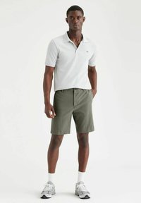 Polo grigio chiaro con maniche corte e colletto, abbinato a pantaloni corti verde oliva. Il modello indossa sneakers grigie con dettagli bianchi.