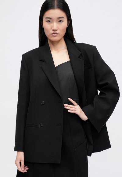 Blazer oversized preto feito de tecido texturizado, com botões duplos, lapelas afiadas e bolsos laterais. Usado sobre uma blusa cinza.