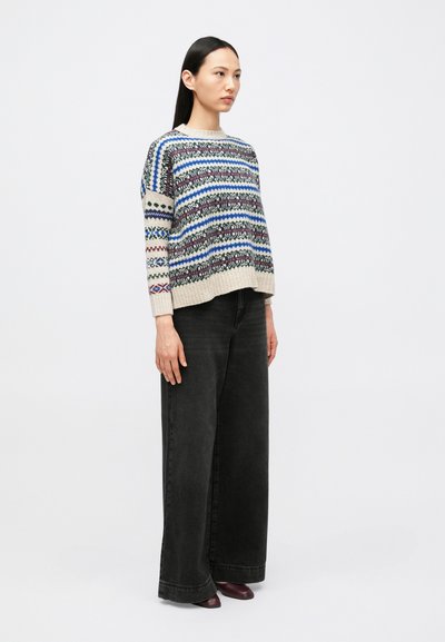 Young woman with long dark hair wearing a patterned knit sweater, wide-leg black jeans, and dark pointed shoes standing sideways.