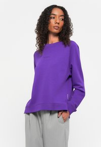 Felpa viola con scollatura a girocollo, caratterizzata da un sottile dettaglio del logo e polsini a costine, abbinata a pantaloni a righe grigio chiaro.