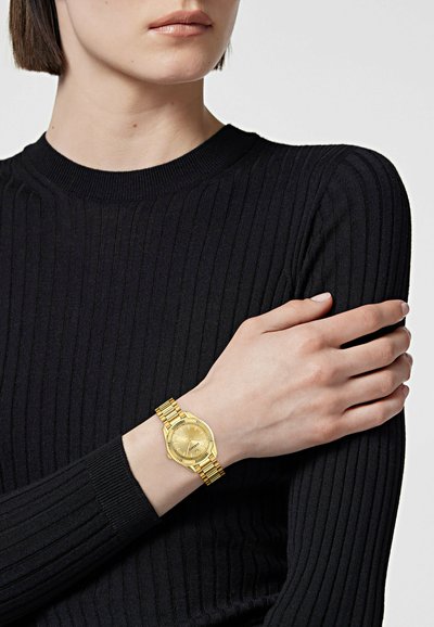Femme portant un haut noir côtelé à manches longues avec une montre en or au poignet gauche, tenant son bras droit à travers son corps.