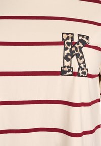 Striped t-shirt in cream with maroon lines; features a leopard-print "K" patch on the left side. Soft fabric, casual design.