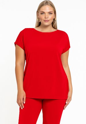 Kvinde med blondt hår iført gyldne øreringe og en klar rød kortærmet top og matchende røde bukser, stående foran en hvid baggrund.