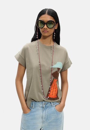 Młoda kobieta o długich ciemnych włosach nosi oversize okrągłe okulary przeciwsłoneczne, beżowy t-shirt z naszywką ptaka z koralików, jasnoniebieskie dżinsy, ręce w kieszeniach.