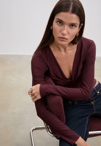 Burgunderfarbener Langarm-Oberteil mit V-Ausschnitt, kombiniert mit dunklen Jeans. Das Model sitzt in einem modernen Stuhl, vor einem sanft beleuchteten Hintergrund.