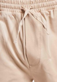 Pantaloni bej cu talie elastica si snur, dotati cu buzunare laterale si o textura a materialului neteda. Materialul pare usor si lejer.