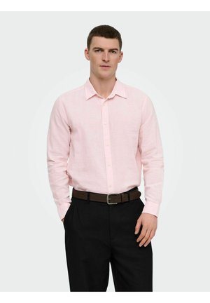 Homme portant une chemise rose clair à manches longues boutonnée, un pantalon noir et une ceinture marron foncé, debout avec une main dans la poche sur un fond uni.