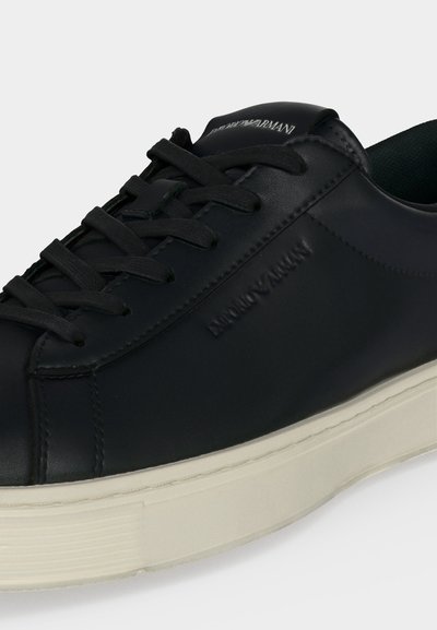Baskets en cuir noir avec lacets noirs et semelle blanche, avec le logo gaufré "Emporio Armani" sur le côté et la languette.