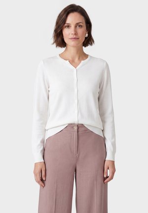 Donna con capelli castani corti che indossa un cardigan bianco abbottonato e pantaloni larghi rosa a vita alta, in piedi su uno sfondo semplice.
