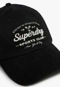 Czarna bawełniana czapka z daszkiem z białym haftowanym napisem "Superdry Sports Club" oraz elementami graficznymi. Regulowany pasek z tyłu.
