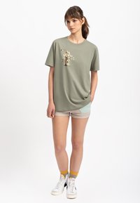 T-shirt in cotone verde oliva con stampa artistica, maniche corte, vestibilità comoda, abbinato a pantaloni corti beige chiaro con una tasca blu.