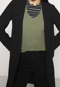 Zwarte blazer over een groen gebreide trui met een V-hals, gecombineerd met zwarte broek met hoge taille; gestreept overhemd zichtbaar onder de trui.