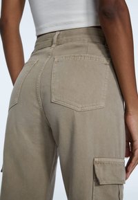 Beige cargobroek met een hoge taille, meerdere zakken en een gestructureerde stof, gezien van de achterkant met de nadruk op gestikte naden en zakontwerp.