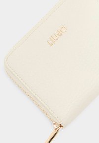 Krémová peněženka na zip vyrobená z texturované umělé kůže, s zlatým logem a kováním zipu pro bezpečné uzavření.