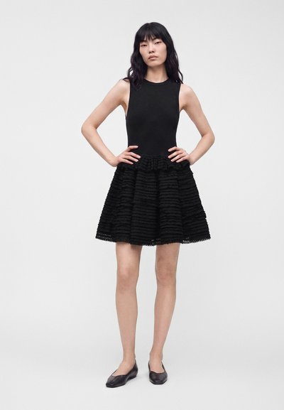 Femme aux longs cheveux noirs, debout, les mains sur les hanches, portant une robe noire sans manches avec une jupe texturée et des ballerines noires.