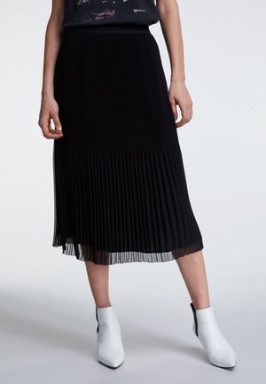 Femme portant une jupe midi plissée noire et des bottines blanches, debout les mains détendues le long du corps, sur un fond uni.