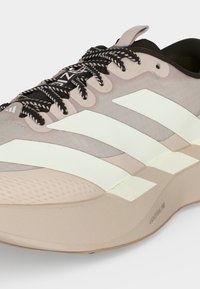 Chaussure de sport beige et blanche avec des lacets noirs et blancs, trois bandes blanches sur le côté, et une semelle épaisse texturée nommée Lightstrike Pro.