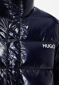 Marineblå pufferjakke med en blank finish, der har quiltet syning og høj krave. Logoet "HUGO" er trykt i hvidt på brystet.