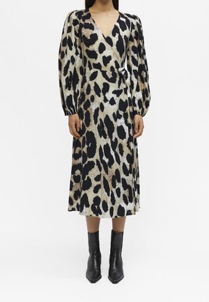 Kobieta ubrana w midi kopertową sukienkę z długim rękawem, dekoltem w serek i panterkowym nadrukiem, do tego czarne botki na białym tle.