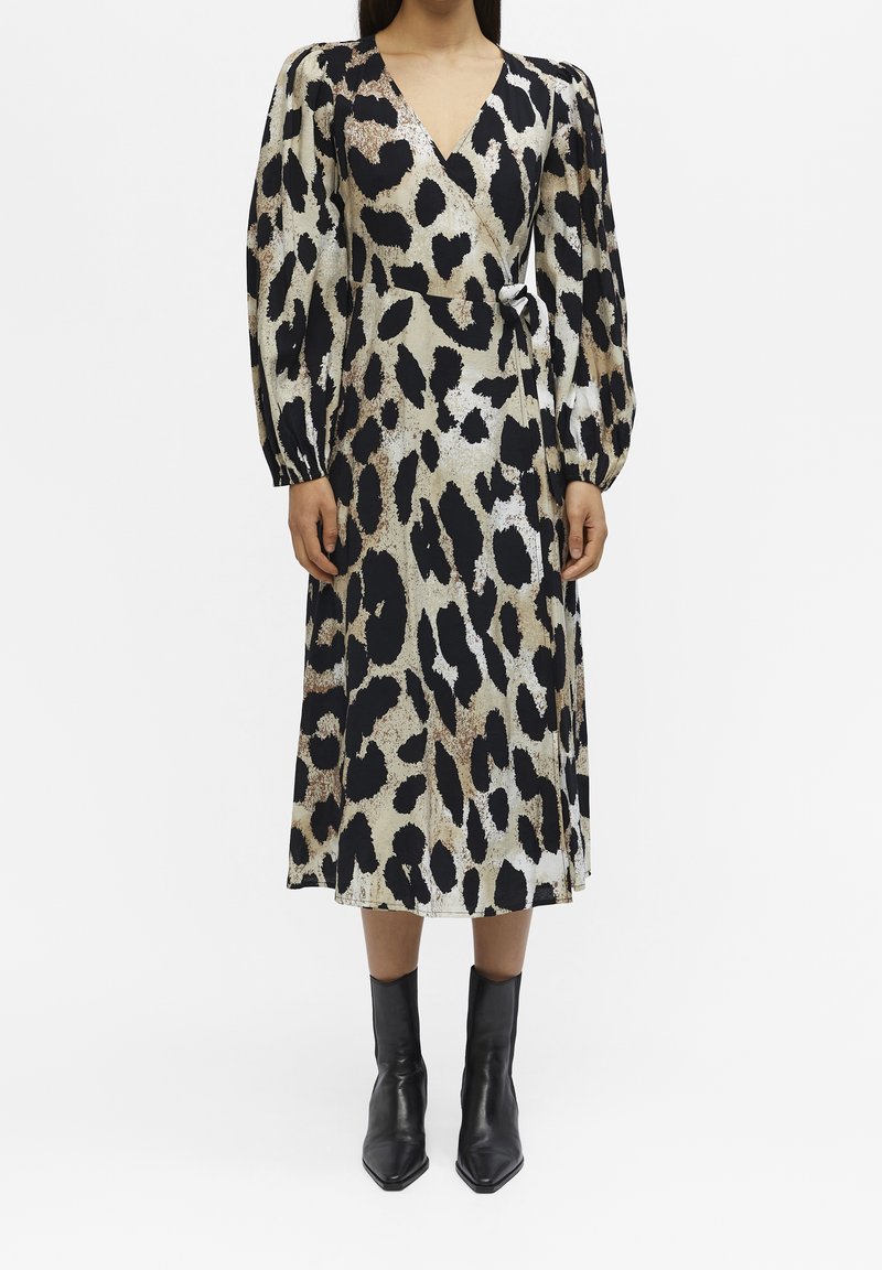 Femme portant une robe midi portefeuille à manches longues, col en V, imprimé léopard, associée à des bottines noires sur fond blanc.