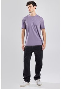 Lyhythihainen laventelit-paita, pyöreä kaula-aukko, rento malli, yhdistettynä väljiin mustiin farkkuihin ja valkoisiin tennareihin mallilla, joka seisoo neutraalilla taustalla.