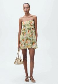 Bloemenminidress in geel met groene en oranje patronen. Heeft dunne spaghettibandjes, een gerimpelde taille en een losse pasvorm, aangevuld met een beige handtas.