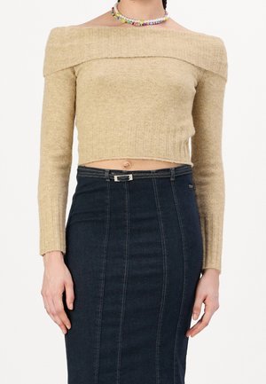 Jersey corto beige con hombros descubiertos y mangas largas, combinado con una falda vaquera oscura de cintura alta y un collar de cuentas colorido.